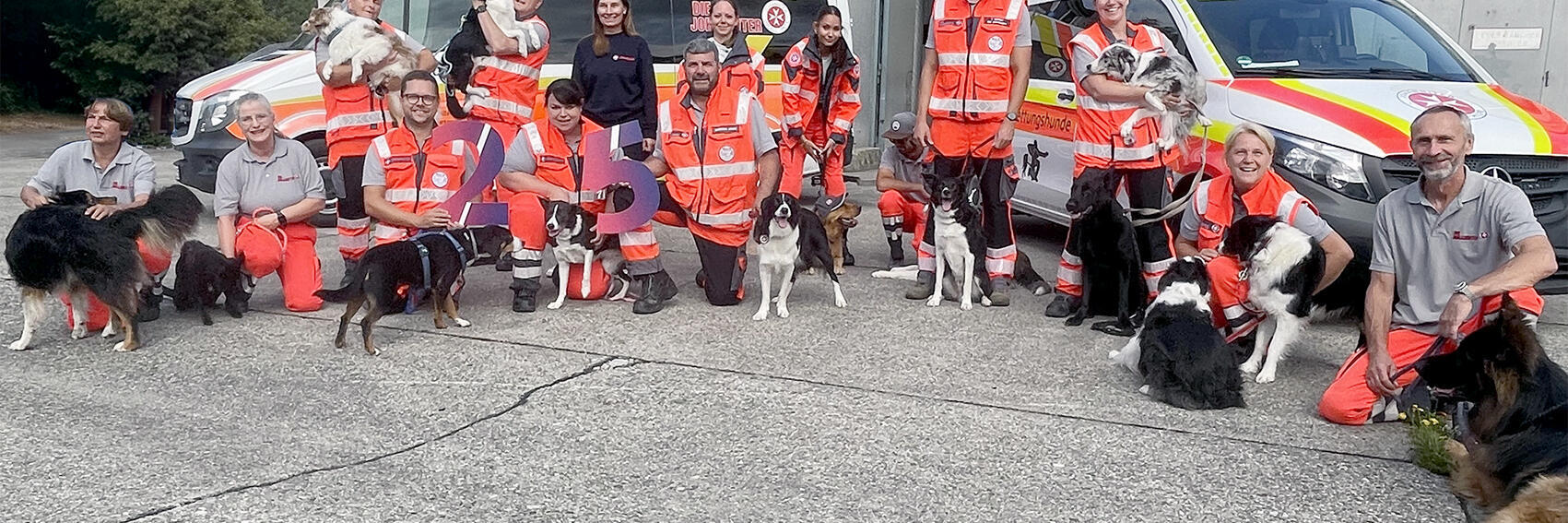 Gruppe von Rettungshundeteams in orange-grauer Einsatzkleidung steht und kniet mit ihren Hunden vor zwei Einsatzfahrzeugen; sie halten große bunte Zahlen „25“.