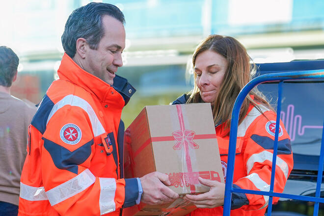 Ein Mann und eine Frau in Johanniter Einsatzkleidung tragen zusammen ein Paket des Johanniter Weihnachtstruckers.