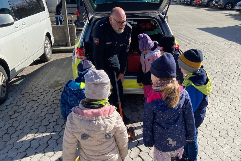 Kinder stehen vor geöffnetem Kofferraum eines Polizeiautos, Polizist spricht zu ihnen.