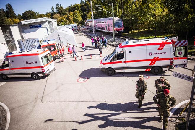 Überblick über ein Einsatzszenario: Zwei Rettungswagen der Johanniter und mehrere Einsatzkräfte stehen auf einer Straße, daneben ein langer roter Zug. Einige Einsatzkräfte sind in orangefarbener Einsatzkleidung, andere in militärischer Uniform zu sehen. Im Hintergrund befinden sich Zelte und Einsatzmaterial.