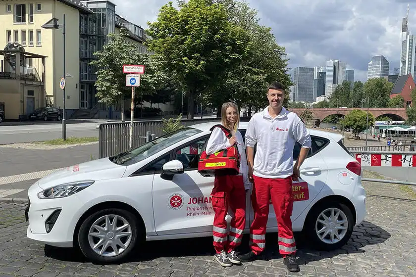 Zwei Freiwilligendienstleistende aus dem Bereich Hausnotruf posieren vor dem Einsatzfahrzeug