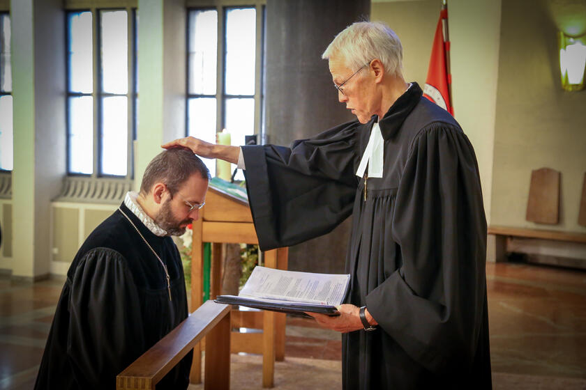 Ein Pfarrer segnet einen knienden Mann in einer Kirche und liest dabei aus einem Dokument.
