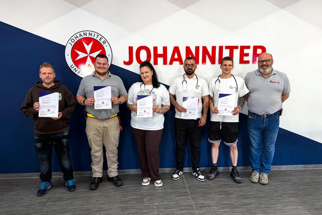 Vier Männer und eine Frau halten ein Zertifikat in der Hand. Neben ihnen steht ein Mann mit Johanniter-Polo. Alle stehen vor einer Wand mit Johanniter-Logo. 