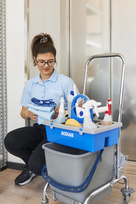 Eine Haureinigungskraft hockt neben ihrem Wagen mit Putz- und Hilfsmitteln.