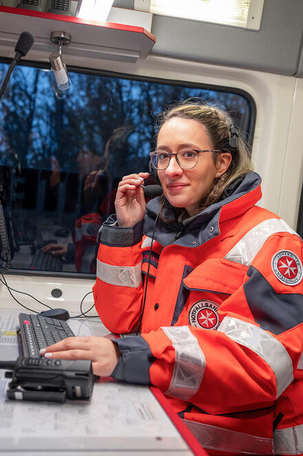 Eine Notfallsanitäterin in einer mobilen Leitstelle sitzt vor Bildschirmen, Knöpfen und Anzeigen und hält das Mikrofon ihres Headsets.