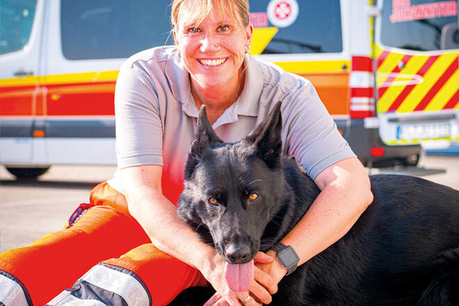 Eine Johanniterin umarmt einen Hund der die Zunge raushängen lässt.