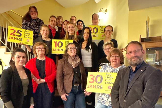 Langjährige Mitarbeitende der Johanniter-Unfall-Hilfe zeigen sich auf einem Gruppenfoto mit Schildern zum 10, 20 und 30 Jährigem Jubiläum. 