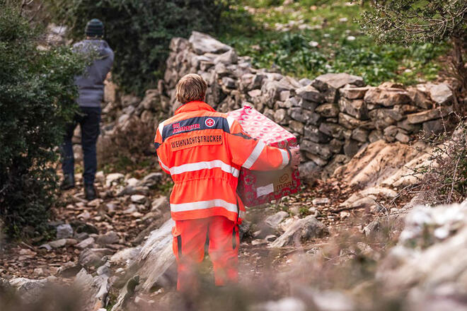Zwei Johanniter tragen Weihnachtstrucker Pakete einen felsigen Weg entlang.