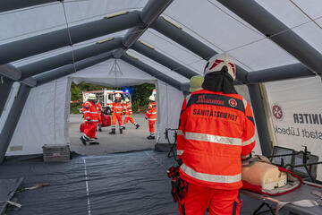 Ein Ehrenamtlicher steht im Behandlungszelt mit dem Rücken zur Kamera. Vor ihm liegt eine Puppe als Darsteller eines Patienten.