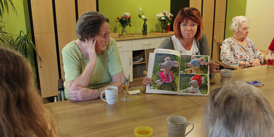 Eine Frau sitzt an einem Tisch mit älteren Damen und zeigt ein Bilderbuch