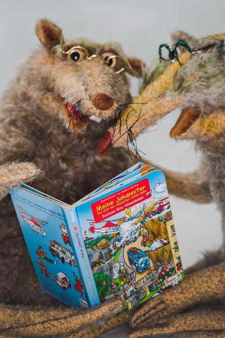Zwei Stoffratten mit Brillen halten gemeinsam das Buch „Meine Johanniter“ in den Pfoten und lesen darin.