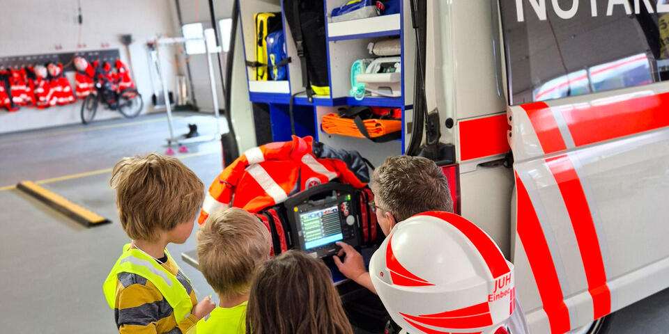 Ein Johanniter erklärt vier Kindern ein medizinisches Gerät. Ein Mädchen hat einen Sicherheitshelm auf