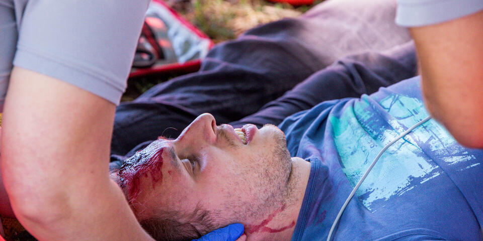 Rettungshelfer*innen stabilisieren im Freien einen gestellten verletzten Mann mit Kopf- und Halsverletzungen und führen Erste‑Hilfe‑Maßnahmen durch.