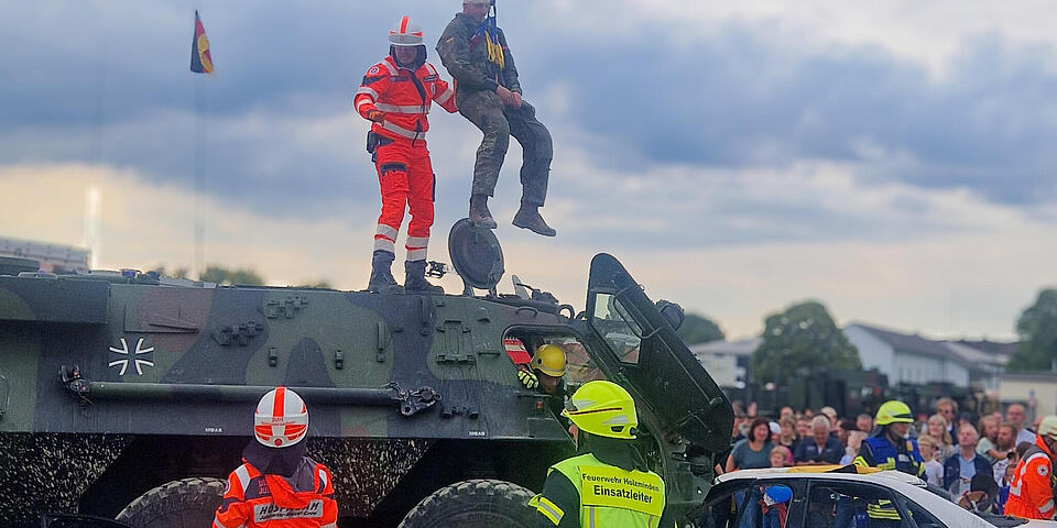 Ein Soldat mit bandagiertem Kopf wird mittels eines Krahns aus einem Truppentransportpanzer, der mit einem PKW kollidiert ist, gezogen. Ein Mann mit Einsatzkleidung und Helm steht auf dem Panzer und unterstützt. Auf dem Boden stehen ein Mann in Johanniter-Einsatzkleidung und Ein Feuerwehrmann mit gelber Wese auf der steht Einsatzleiter. Im Hintergrund schauen Menschen zu.