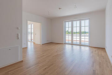 Leerer, heller Wohnraum mit Holzfußboden, großen Fenstern und Zugang zu einem weiteren Zimmer.