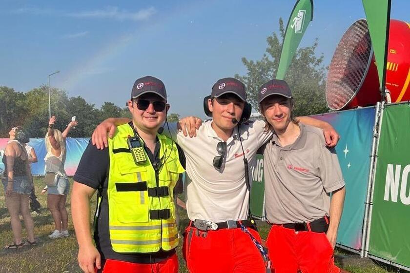 Drei Johanniter-Helfende in Einsatzkleidung stehen Arm in Arm vor einem Festivalzaun; im Hintergrund ein Ventilator, Festivalbesucher*innen und ein leichter Regenbogen.