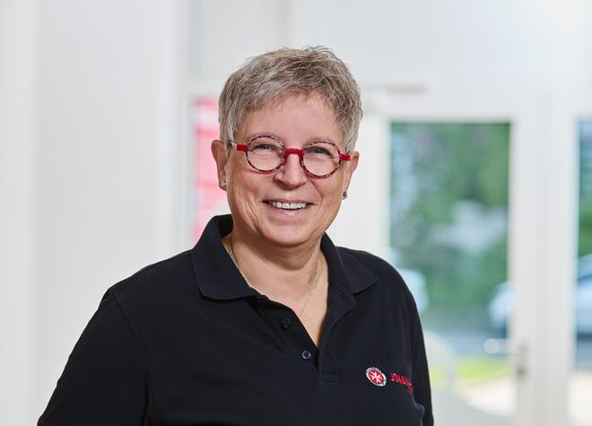 Eine Frau mit grauen Haaren und Brille in einem Eingangsbereich. Sie trägt ein blaues Polo-Shirt