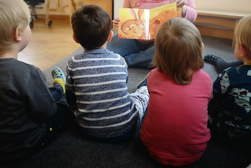 Kinder schauen sich gemeinsam mit der Erzieherin ein Bild zum Thema Herbst an. 