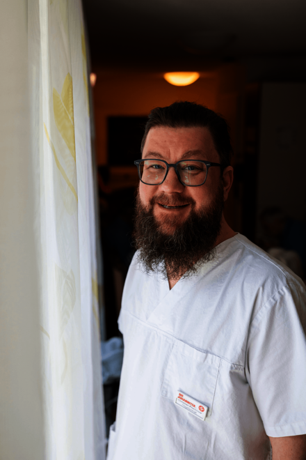 Ein Mann in weißer Arbeitskleidung steht am Fenster neben einem Vorhang, lächelt 