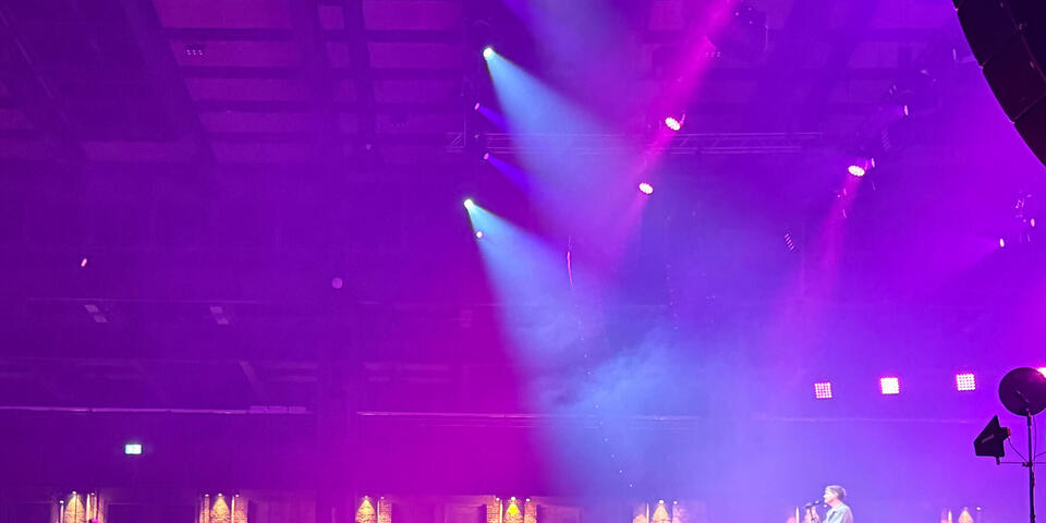 Zuschauer schauen auf eine Bühne. Dort steht ein Mann. In der Luft pinke und blaue Lichteffekte