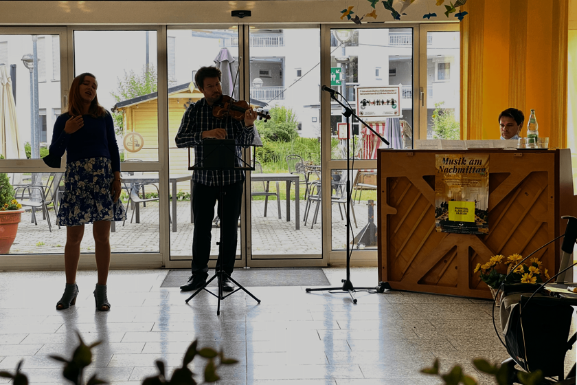 eine Sängerin in geblümtem Rock, eine Geigenspieler in einem karierten Hemd und ein Mann am Klavier musuzieren