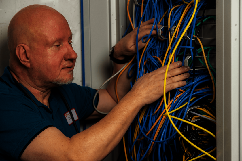 Ein kahlköpfiger Mann mit weißem Kinnbart vor einem Schrank mit vielen verschiedenfarbigen Kabeln