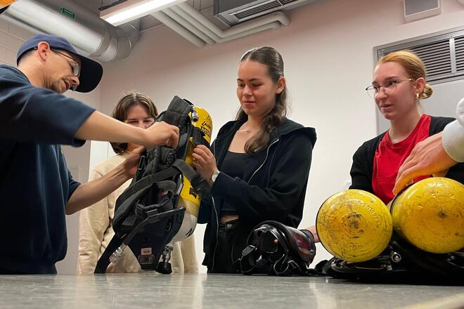 Ein Feuerwehrmann hilft einer Jugendlichen beim Aufsetzen eines Pressluft-Flaschen-Rucksacks während andere Jugendliche gespannt zusehen.