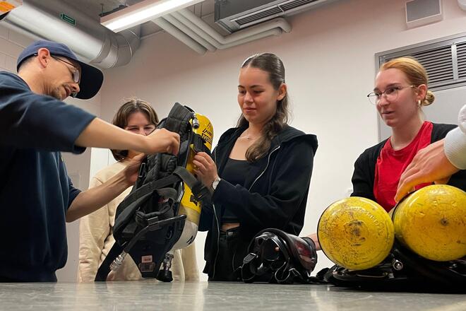Ein Feuerwehrmann hilft einer Jugendlichen beim Aufsetzen eines Pressluft-Flaschen-Rucksacks während andere Jugendliche gespannt zusehen.