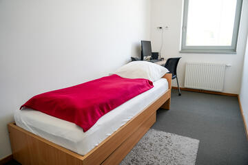 Ruheraum mit Holzbett, roter Decke, weißem Kissen, Arbeitstisch mit Computer und und kleinem Teppich.