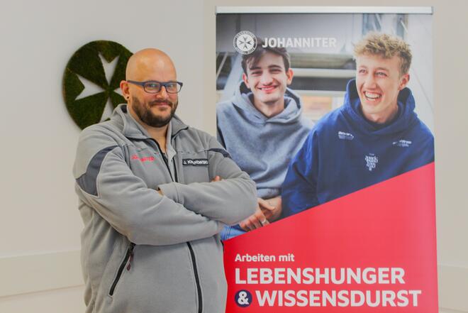 Ein Mann mit Glatze und Bart und mit grauer Fleecejacke vor einem Roll-Up auf dem zwei junge Männer zu sehen sind. Auf dem Roll-Up steht "Arbeiten mit Lebenshunger und Wissensdurst"