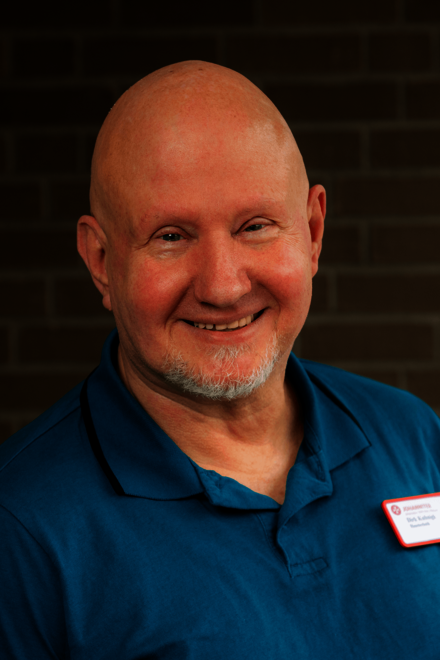 Kopf und Schultern eines kahlköpfigen Mannes mit weißem Kinnbart in einem Poloshirt in Türkis. Er steht vor einer gemauerten Wand und lächelt in die Kamera.