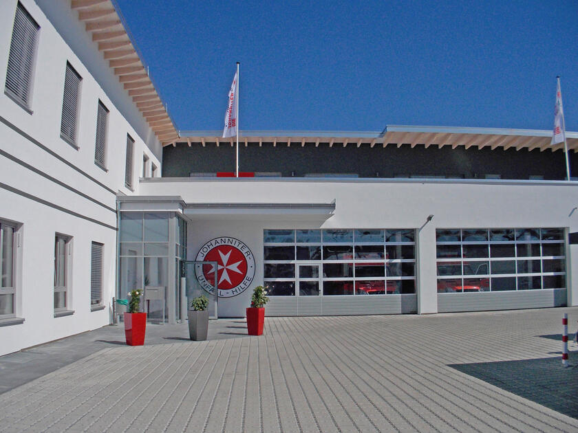 Auf dem Bild ist die Regionalgeschäftsstelle der Johanniter in Nürnberg zu sehen. Es ist ein L-förmiger Bau mit einem großen Logo am Eingangsbereich. Auf dem Dach wehen Fahnen.