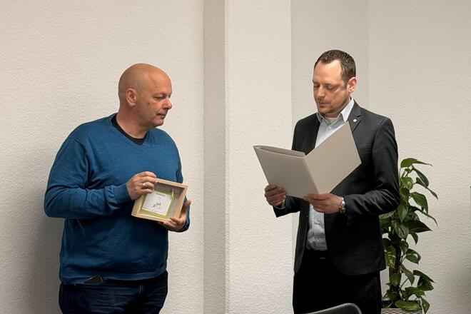 Ein Mann hält ein Geschenk in der Hand. Ein anderer hält eine Urkunde in der Hand und lies sie vor.