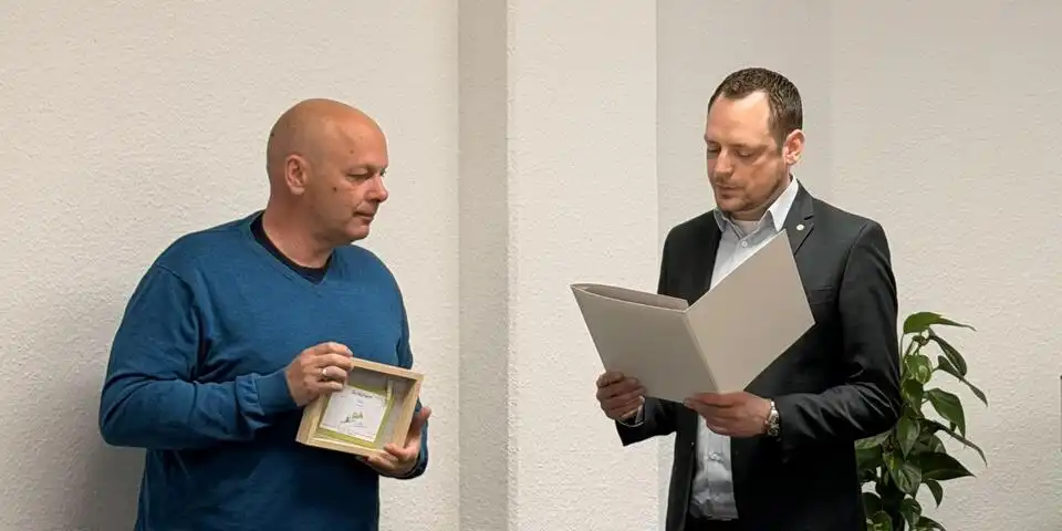 Ein Mann hält ein Geschenk in der Hand. Ein anderer hält eine Urkunde in der Hand und lies sie vor.