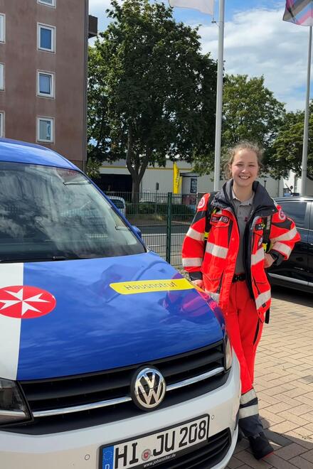 Eine junge Frau in Einsatzbekleidung und mit einem Notfallrucksack steht neben einem Fahrzeug mit Johanniter-Logo auf einem Parkplatz