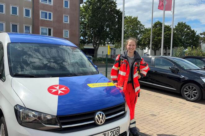 Eine junge Frau in Einsatzbekleidung und mit einem Notfallrucksack steht neben einem Fahrzeug mit Johanniter-Logo auf einem Parkplatz