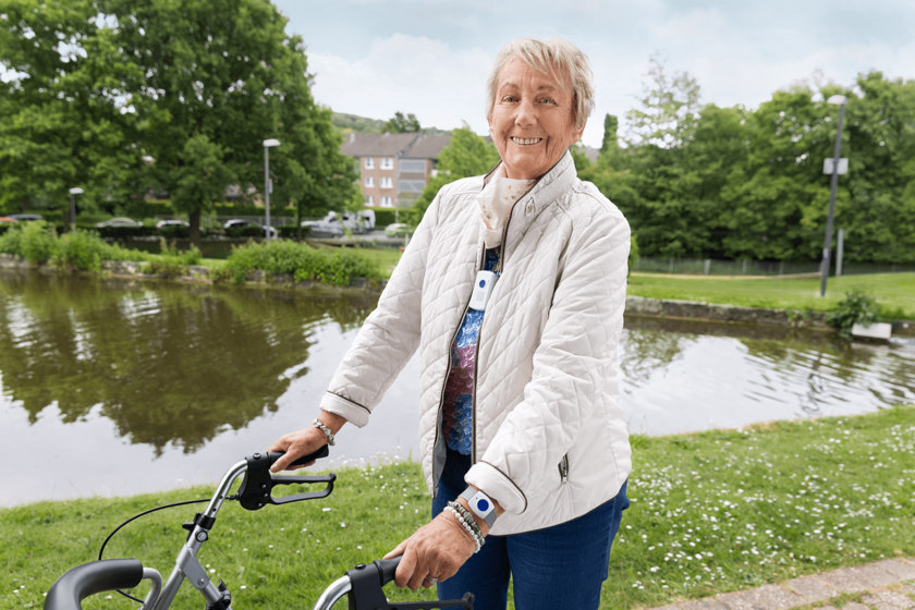 Lächelnde ältere Dame in weißer Steppjacke und Rollator in einem Parkt, im Hintergrund ein See. Um den Hals und am Handgelenk trägt sie einen Notrufknopf