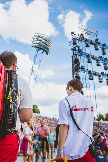 Ehrenamtliche Johanniter-Sanitäter bei einem Großevent. Sie tragen Einsatzkleidung und stehen inmitten einer Menschenmenge vor einer Bühne mit Flutlichtanlagen und einem blauen Himmel.