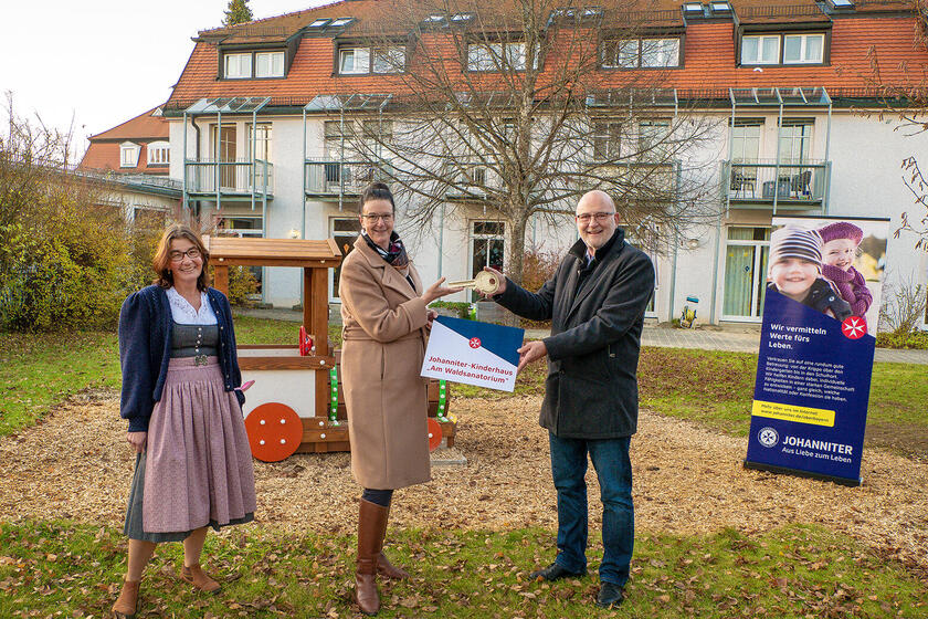 Drei Erwachsene stehen vor dem Johanniter-Kinderhaus „Am Waldsanatorium“, während ein Mann einer Frau symbolisch einen großen Schlüssel für die Einrichtung überreicht.