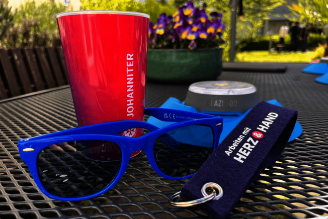 Roter Becher, blaue Sonnenbrille und Schlüsselband auf Metalltisch.