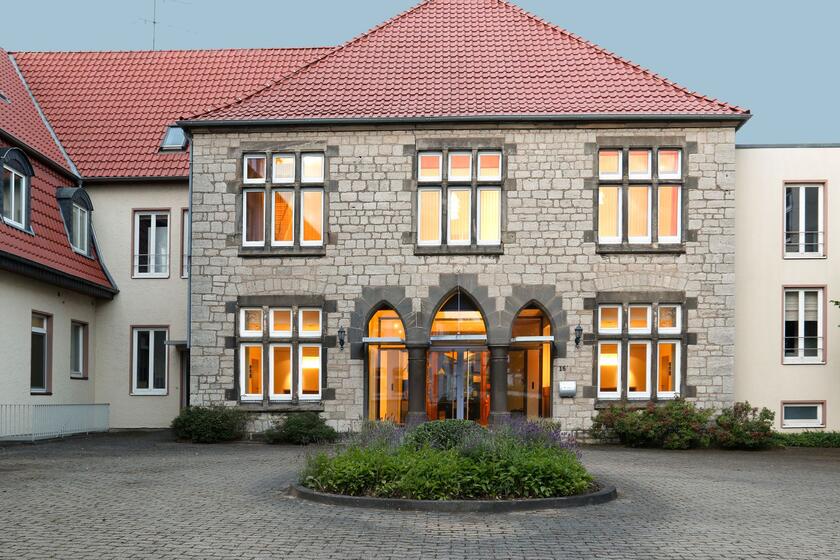 Ein steingebäude mit modernen Erweiterungen links und rechts. davor eine gepflasterte Einfahrt. In deren Mitte eine runde Bepflanzung ist. 