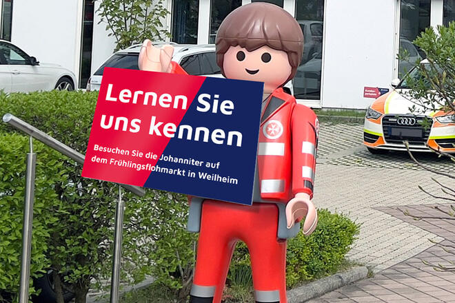 Ein großes Playmobilmännchen in Einsatzkleidung steht auf einem Johanniter Dienststellen Parkplatz und hält ein „Lernen Sie uns kennen“ Schild in der Hand.