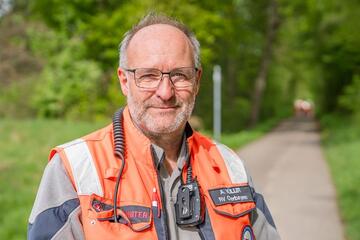 Johanniter im orangefarbenen Einsatzwesten steht im Grünen und blickt lächelnd in die Kamera.