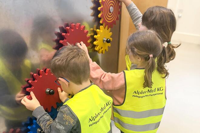 Drei Kinder mit gelben Warnwesten an einer Metallwand an der Zahnräder zum Drehen sind.