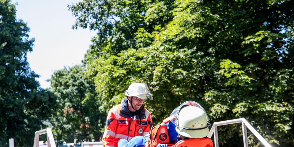 Rettungshelfer*innen in Schutzkleidung tragen auf einer Trage eine verletzte Person und arbeiten konzentriert bei einer Übung im Freien.