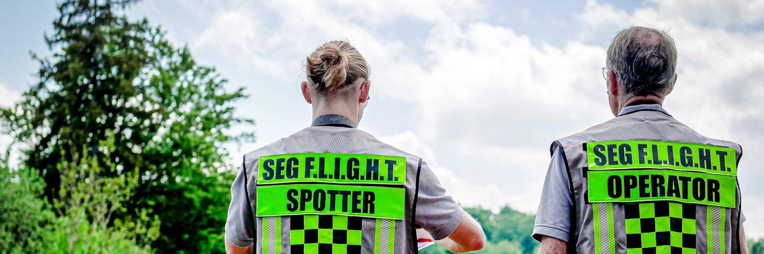 Zwei Johanniter mit Warnwesten, beschriftet mit ‚Spotter‘ und ‚Operator‘, stehen nebeneinander und beobachten eine Drohne, die über ihnen am Himmel fliegt.