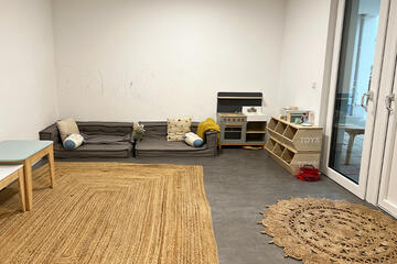 Heller Raum mit Terassentüren und Sisalteppichen. Kleine Tische an der Wand und eine Spielcouch zum Ausruhen. Spielküche aus Holz und ein Holzregal mit Zubehör für die Küche