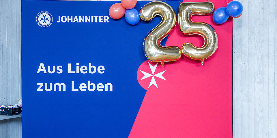 blau-rote Wand mit Johanniter-Logo und Spuch "Aus Liebe zum Leben", davor sind zwei goldene Folienballons mit den Zahlen 2 und 5 zu sehen, die für das 25jährige Jubiläum der Veranstaltung stehen