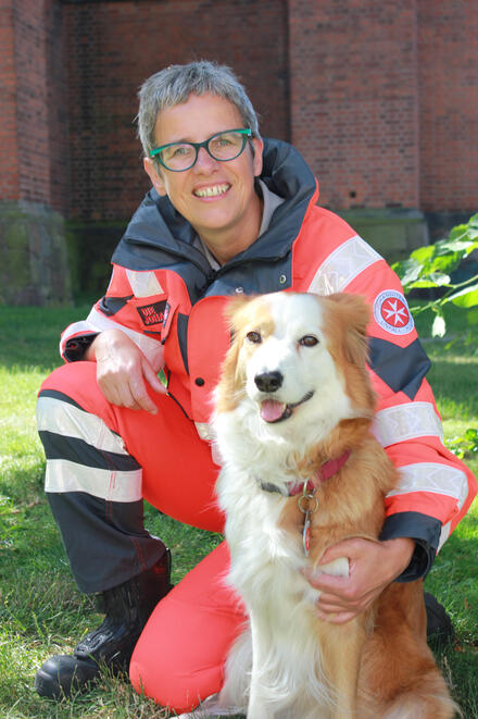 Die Johanniter-Hundeführerin Jana Simon kniet neben ihrem Hund Maja. Die Hündin ist ein Border-Collie in den Farben weiß und braun. Beide lächeln in die Kamera.