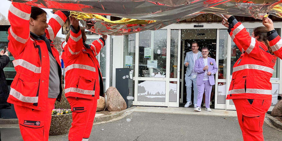 Johanniter in Einsatzbekleidung halten Rettungsdecken hoch. Im Hintergrund kommt ein Paar durch eine Tür